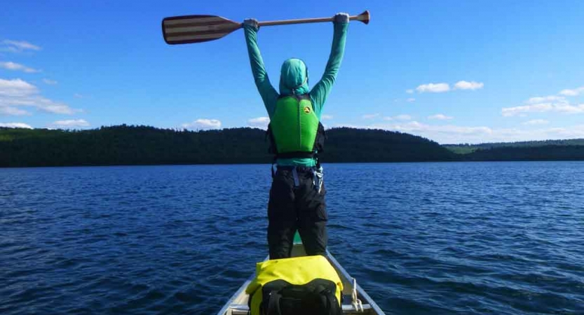 a student stands at the front a canoe and raises their paddle in the air on an outward bound course